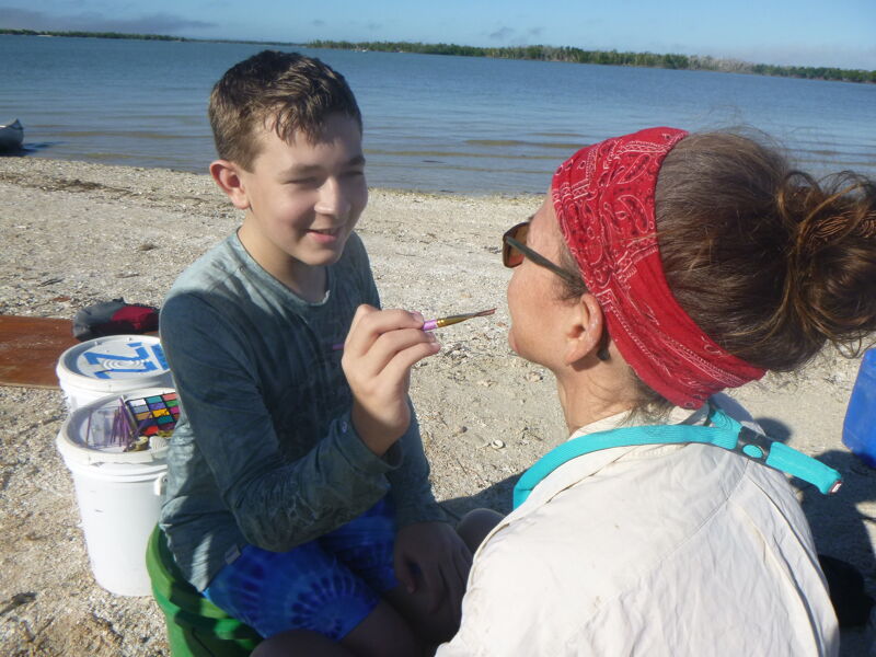 A young boy is carefully applying makeup to a woman's face on a sunny beach. The woman is wearing a red bandana and sunglasses, while the boy is focused on his task. They are surrounded by buckets and other items, suggesting they may be preparing for an event or activity. The background shows a calm body of water and a clear sky.